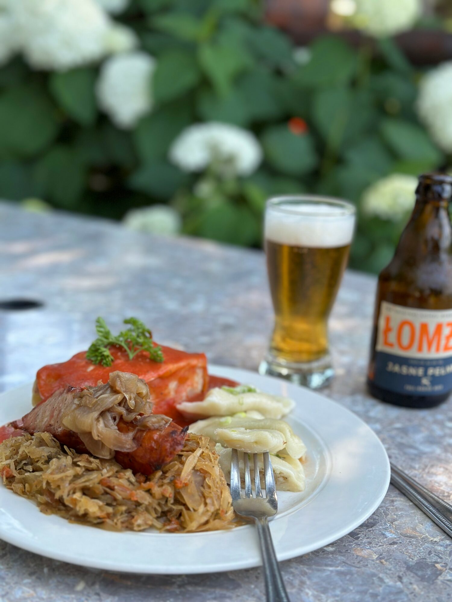 Plate with meat, vegetables, and tomato, a fork, a glass of beer, and a bottle on a stone table outdoors.