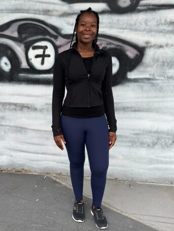 Woman standing outdoors in front of a graffiti wall, smiling, wearing a black jacket, blue pants, and sneakers.