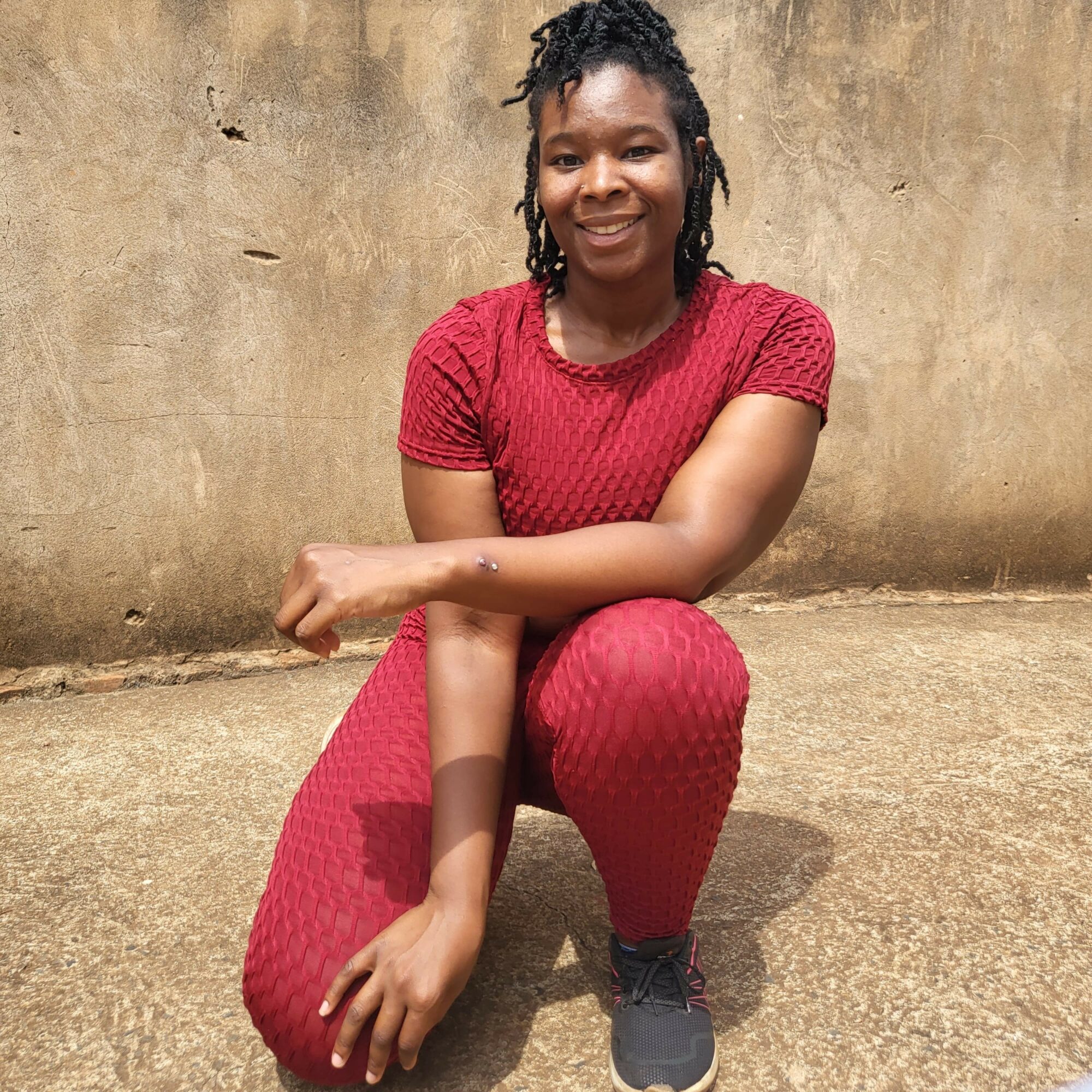Young woman crouching outdoors, smiling, wearing a red textured outfit and black shoes, against a beige wall.