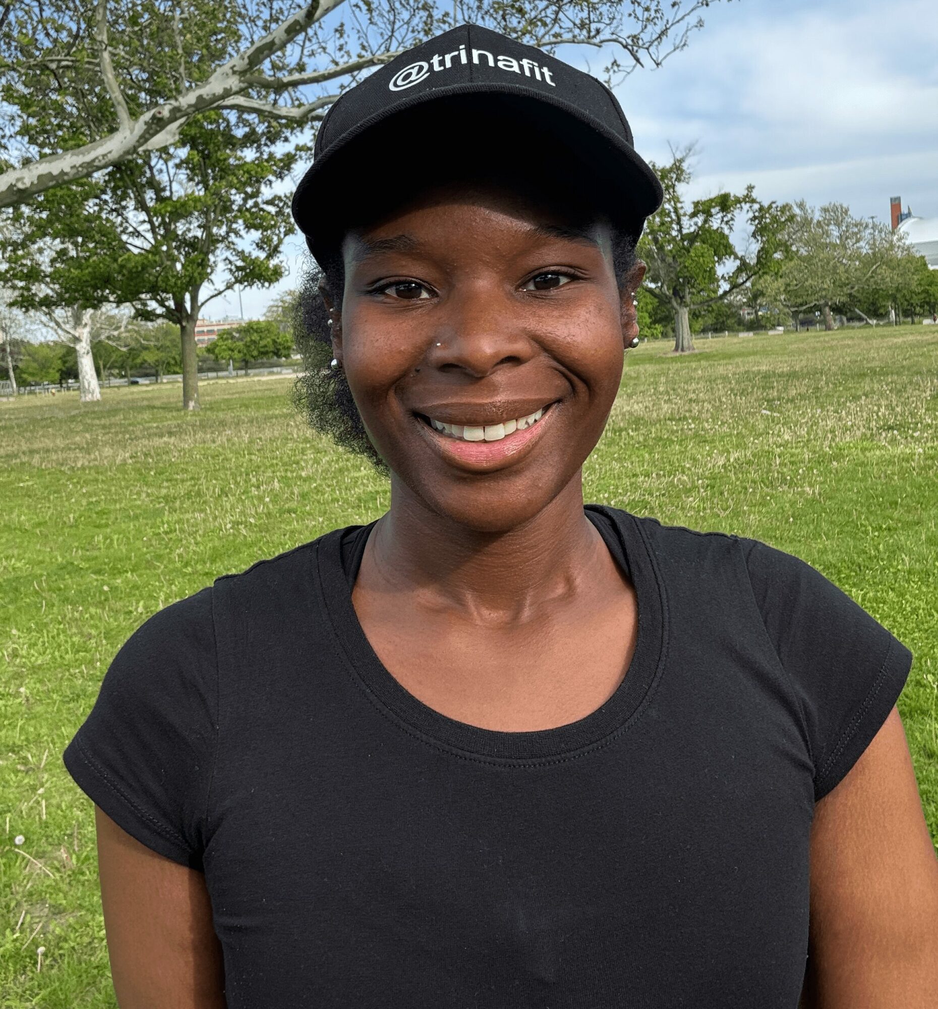 Smiling woman outdoors wearing a black cap and t-shirt, trees and grass in background.