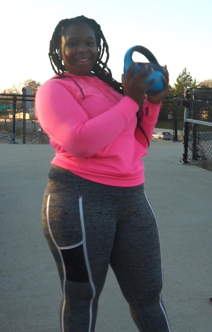 Woman in pink hoodie holding a kettlebell outdoors, with a fence and trees in the background.
