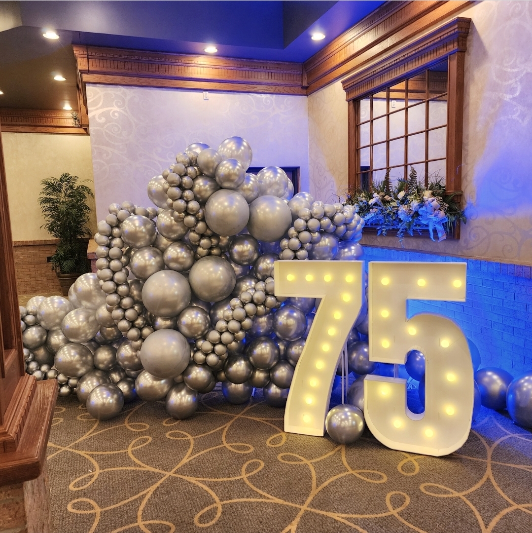 Decorative display with silver balloons and illuminated number 75 in a room with wooden accents and patterned carpet.