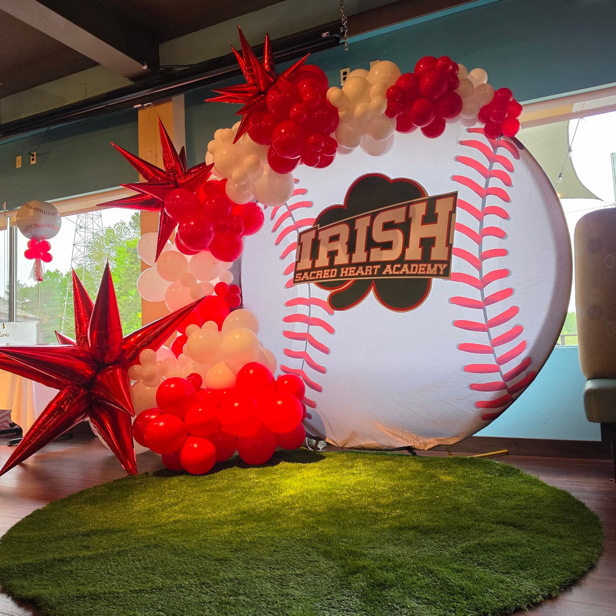 Decorative baseball-themed display with red and white balloons, large red stars, and a logo for Irish Sacred Heart Academy, on a green rug.