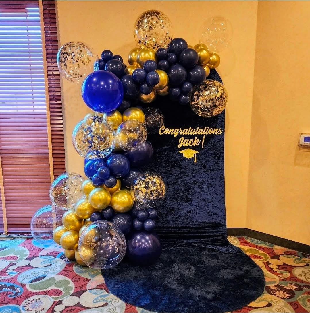 Balloon arrangement with blue, gold, and transparent balloons, and a congratulatory sign on a black backdrop, in a decorated room.