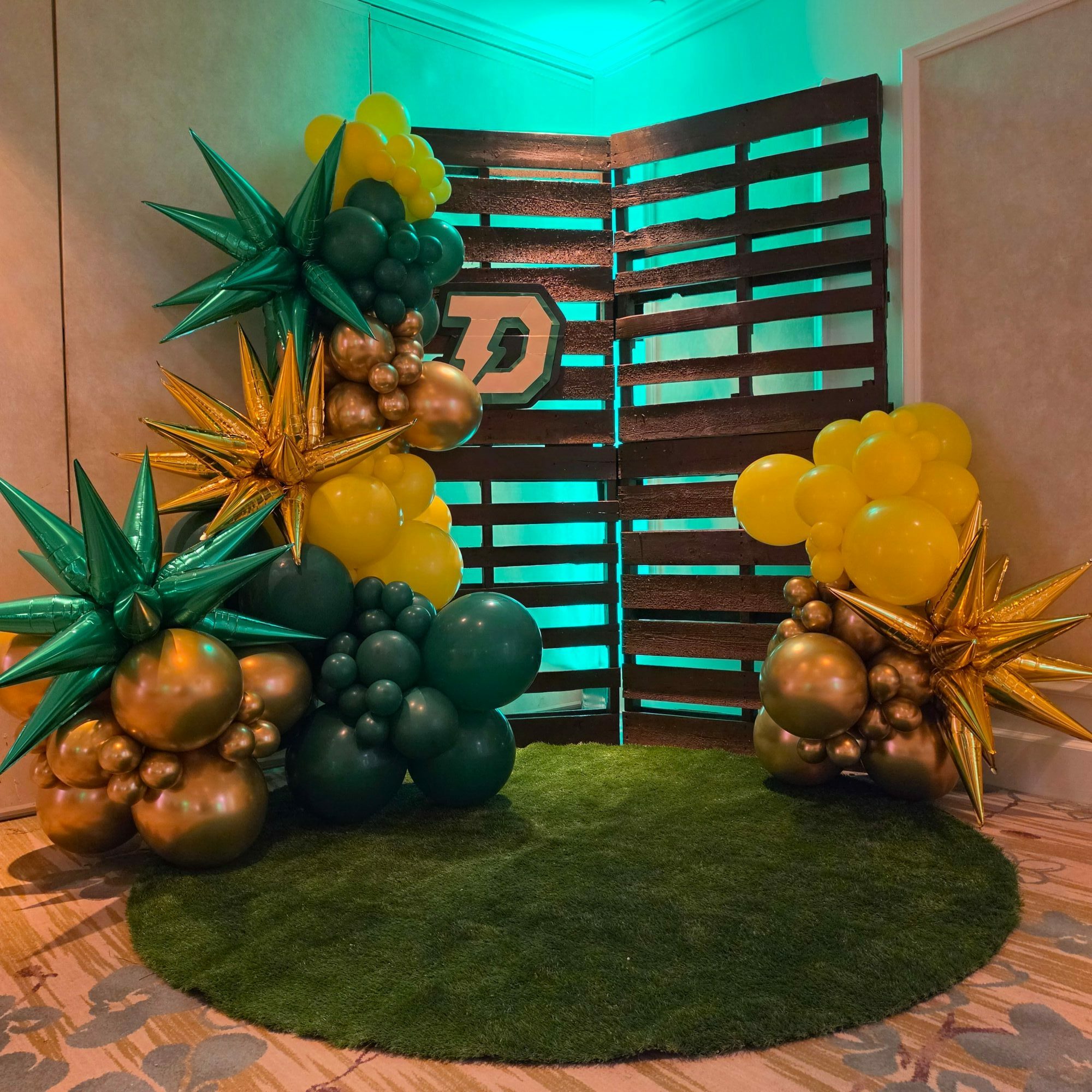 Decorative balloons and metallic ornaments arranged on a green circular rug, with a wooden slatted backdrop and patterned floor.