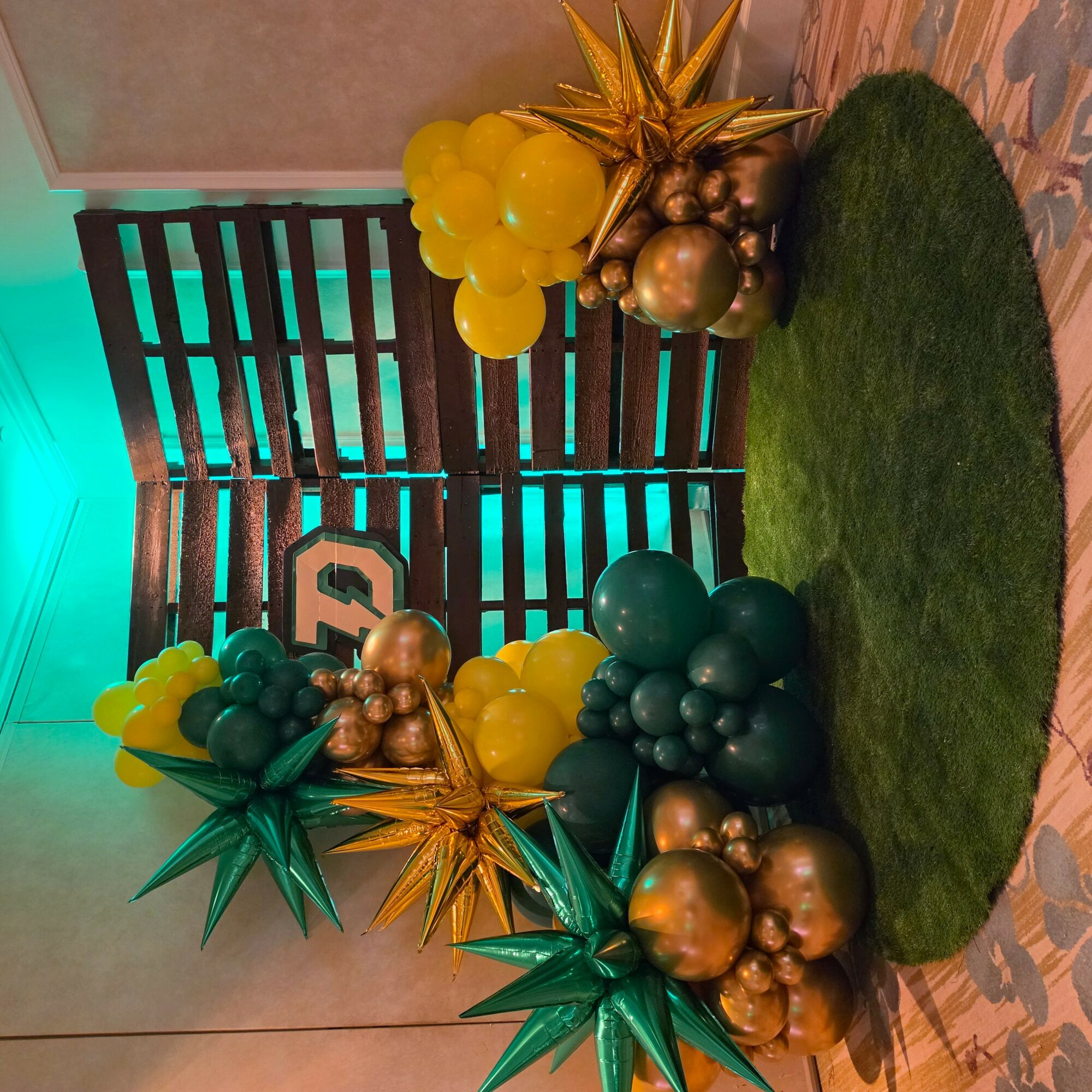 Decorative balloons and metallic ornaments arranged on a green circular rug, with a wooden slatted backdrop and patterned floor.