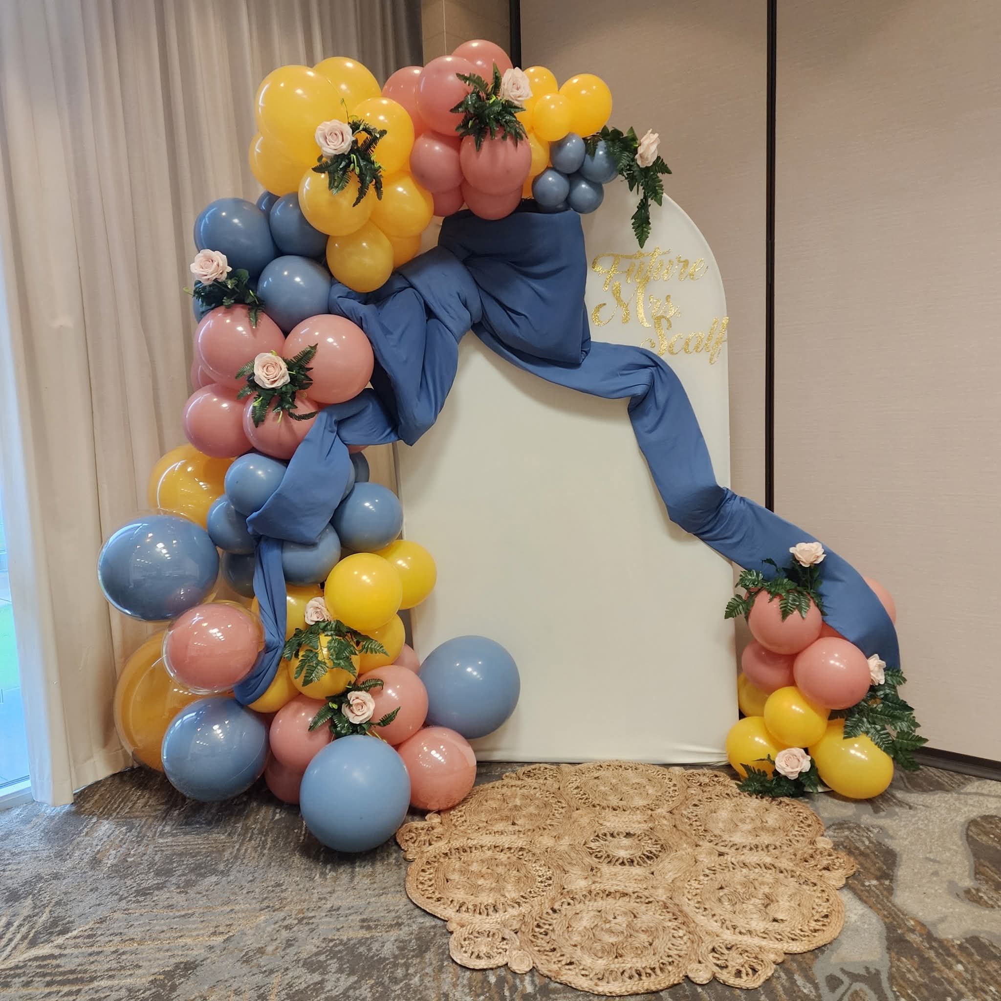 Decorative arch with balloons, flowers, and a blue fabric drape, set against a white backdrop on a carpeted floor.