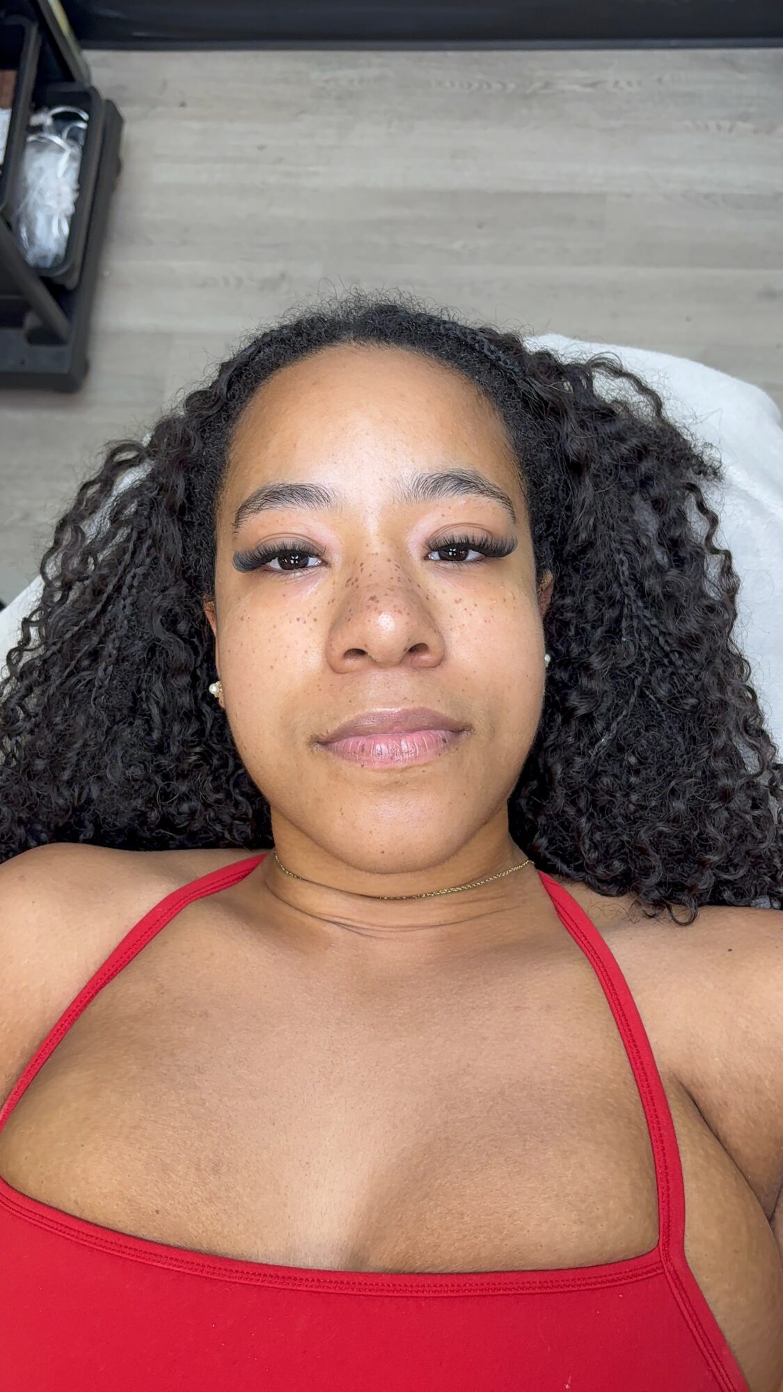 A woman with curly hair lying on a bed, wearing a red top, looking at the camera.