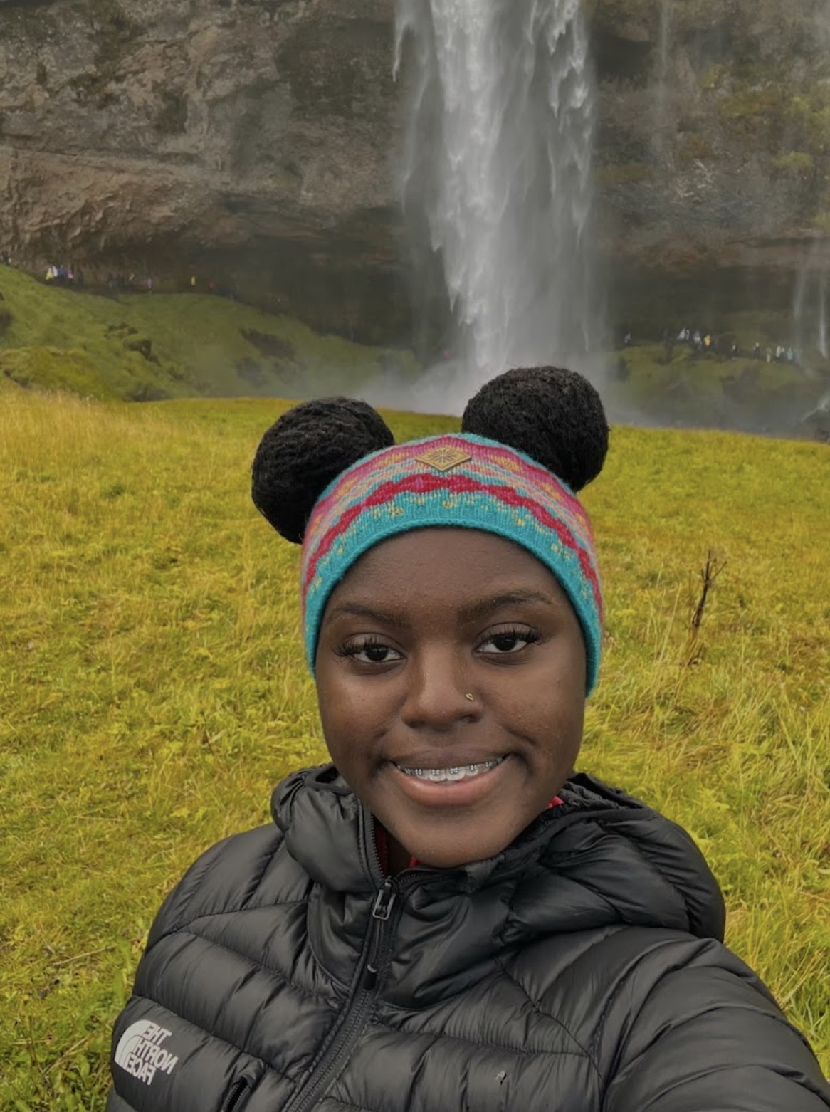 Young girl outdoors in front of a waterfall, wearing a colorful beanie and black jacket, smiling at the camera.