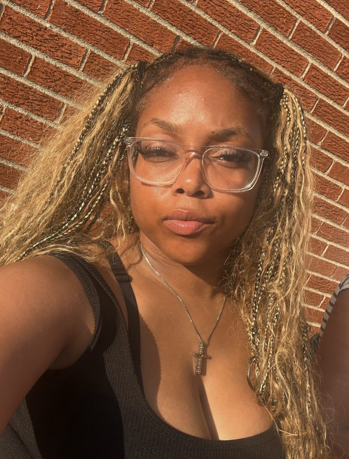 Woman with glasses and long curly hair outdoors against a brick wall, sunlight on face, wearing a black top and cross necklace.