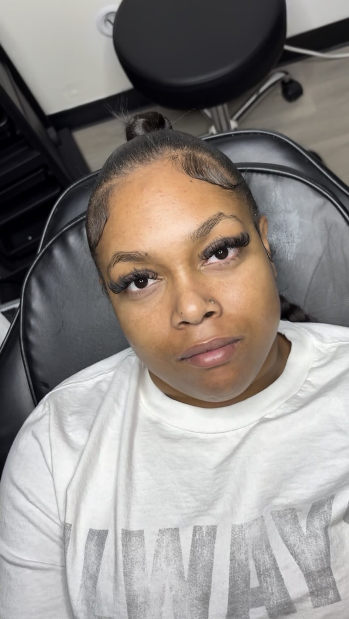 Woman with styled hair lying on a chair, looking at the camera, in a room with a black stool and white wall.
