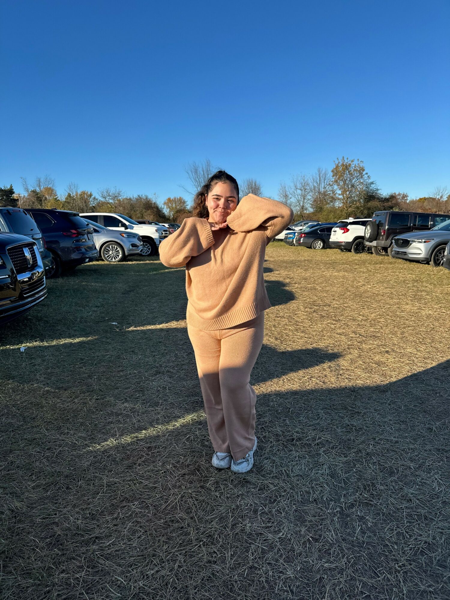 Person standing on grass field, wearing a beige coat and pants, with cars parked in background under clear blue sky.