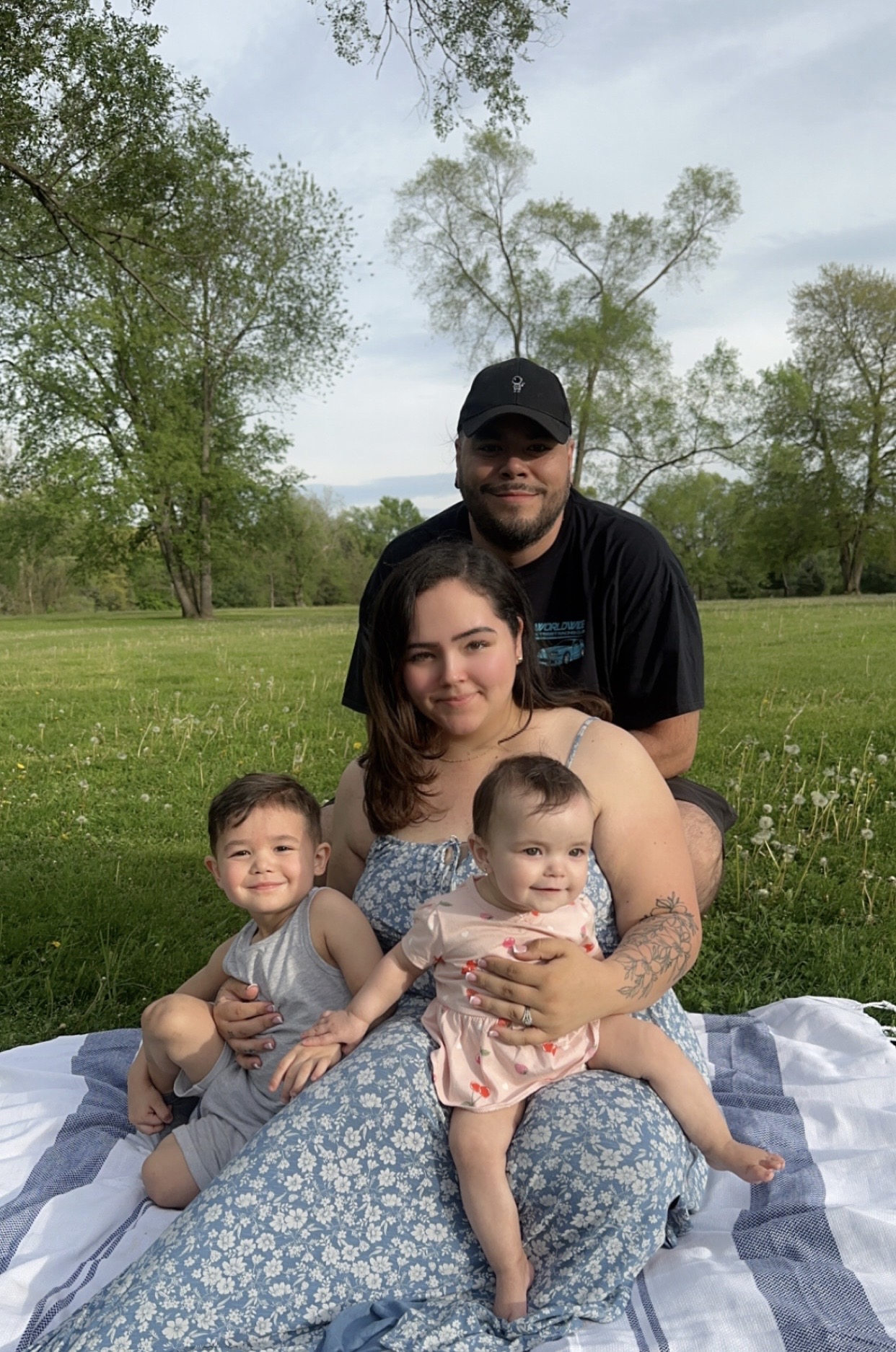Family of four sitting on a blanket outdoors with trees and sky in background.