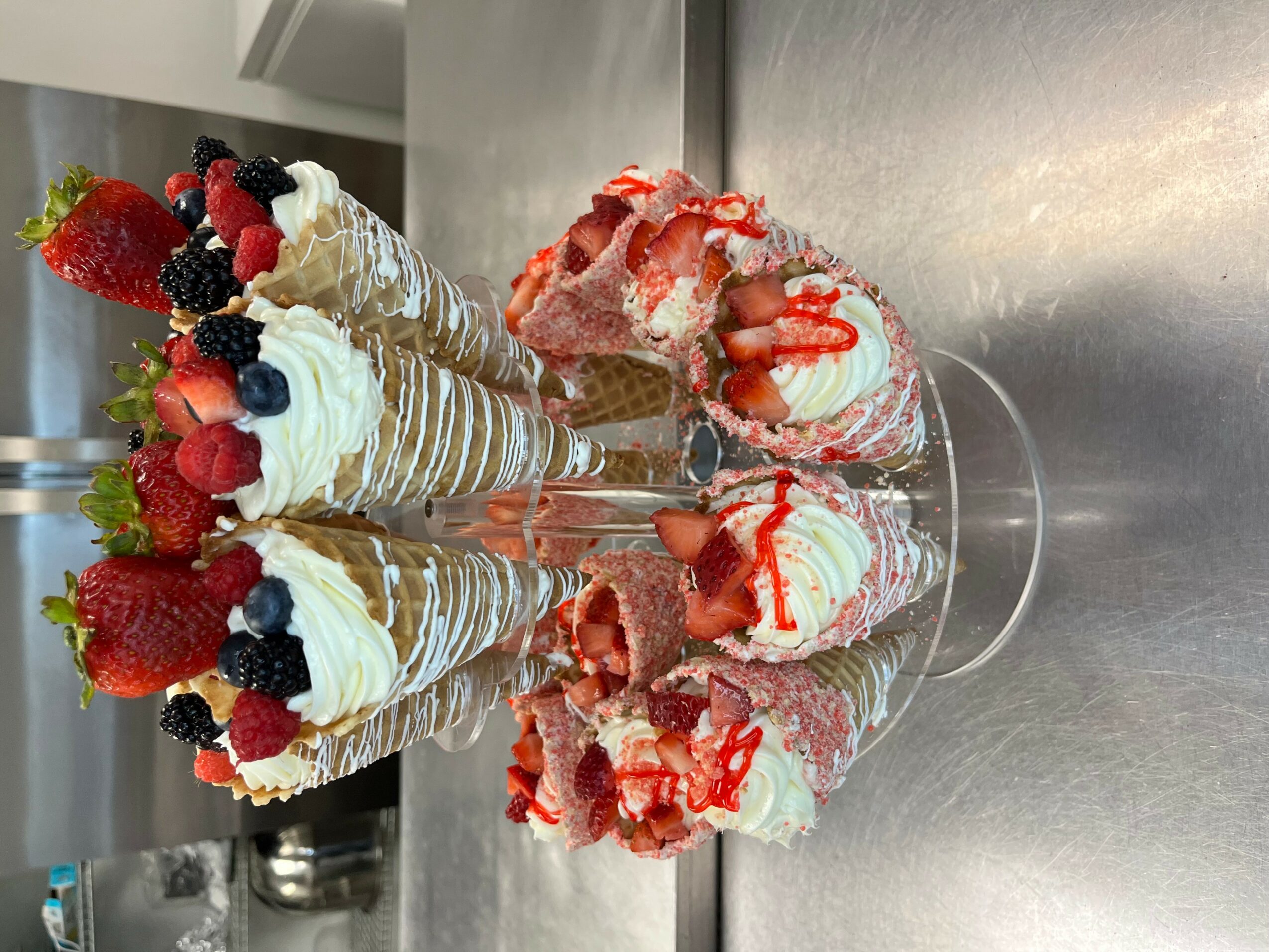 Two tall ice cream sundaes with strawberries, blackberries, and whipped cream in glass cups on a reflective surface.