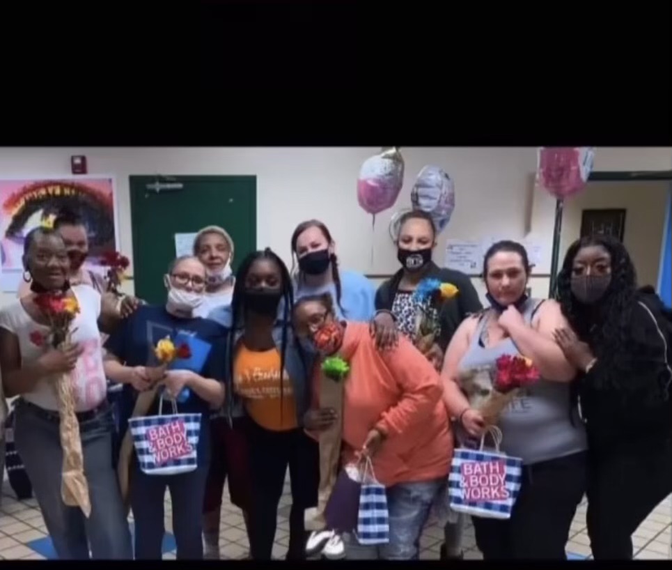 Group of women holding flowers and gift bags, some wearing masks, in a decorated indoor space.