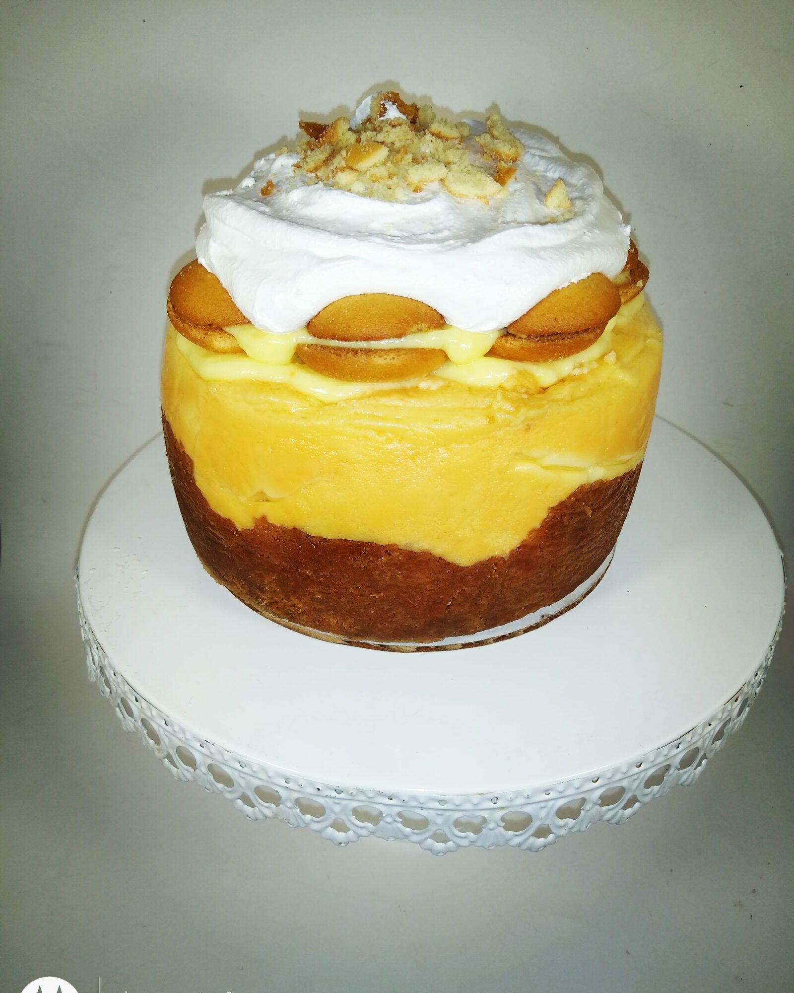 Layered dessert with yellow custard, topped with whipped cream, peach slices, and nuts on a white doily-covered surface.