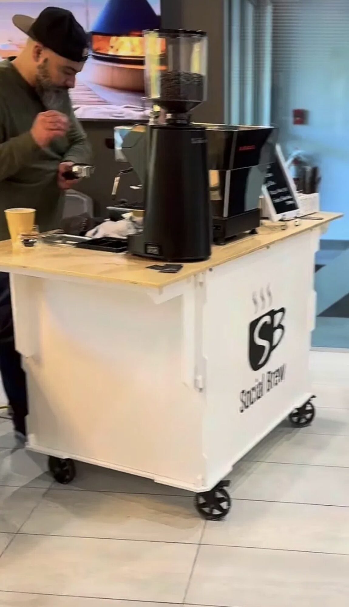 Person in a black hat and green shirt at a coffee cart with coffee machine, cups, and a window in background.