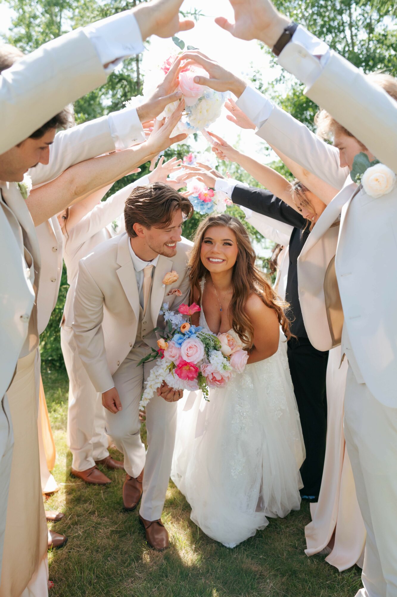 Bride and groom smiling under a heart-shaped arch formed by friends' raised arms outdoors, holding a bouquet.
