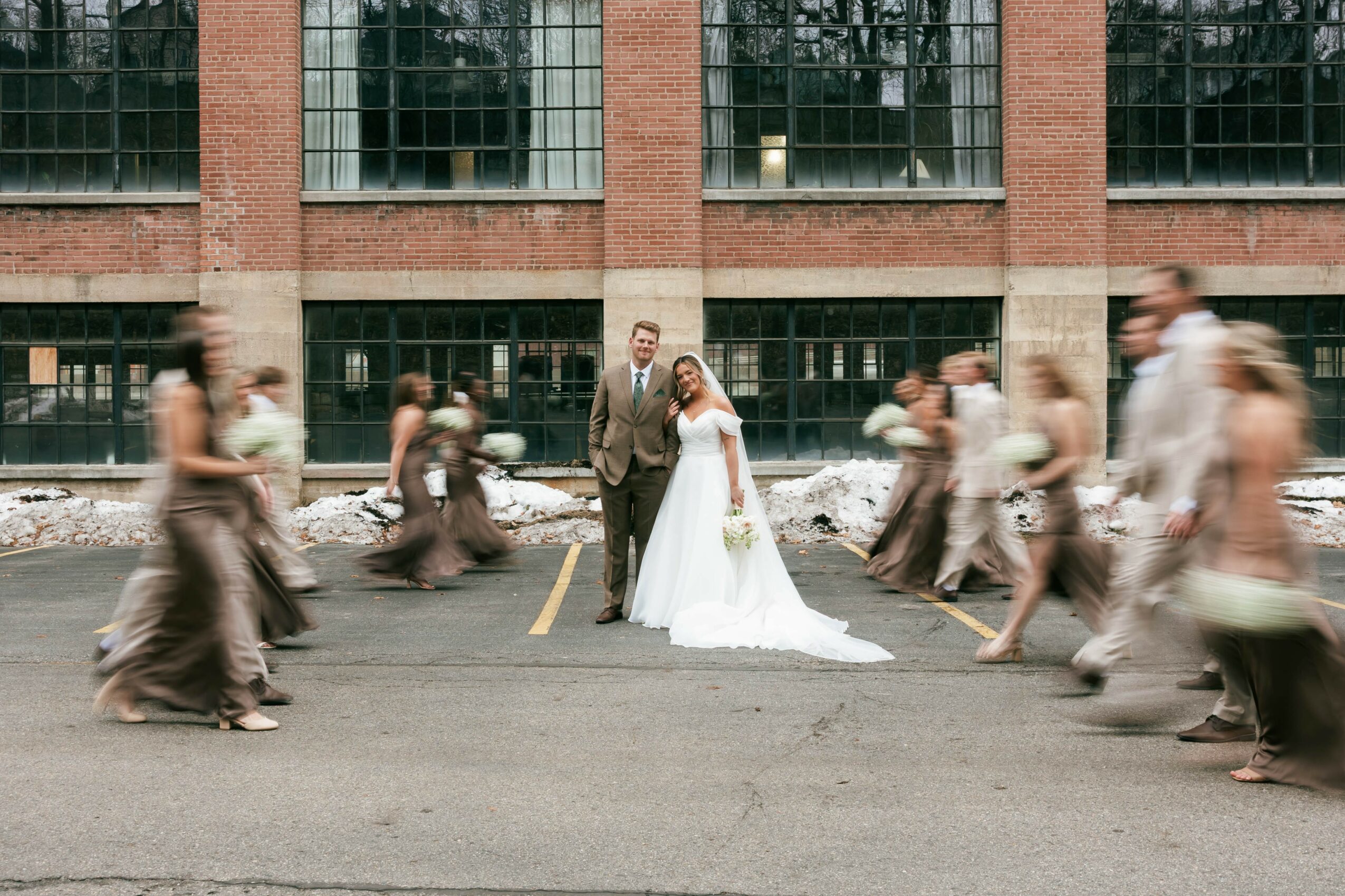 A bride and groom stand in the center of a street, surrounded by blurred people walking past, in front of a brick building.