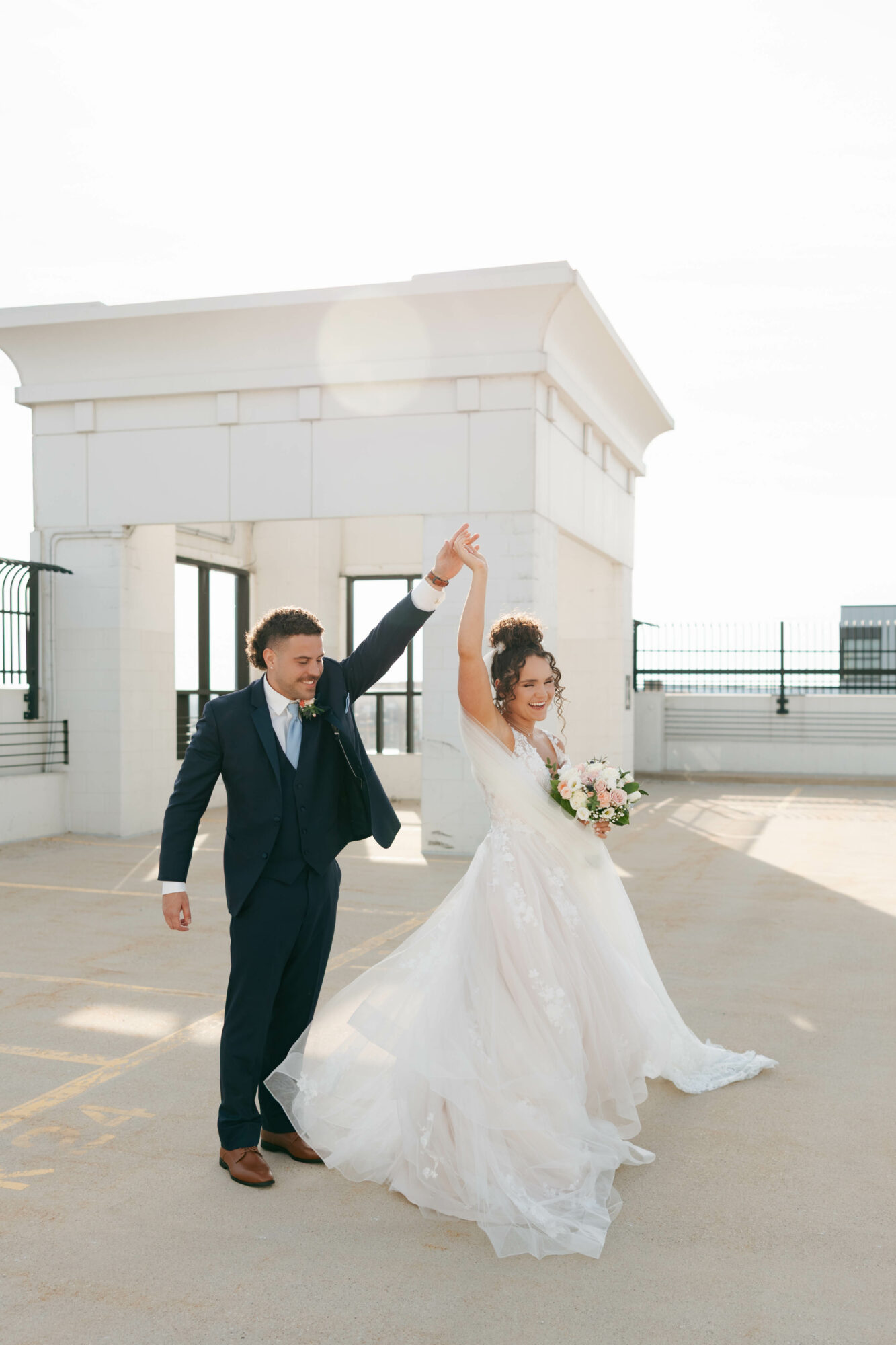 A bride and groom dance outdoors near a white structure, holding hands up, with a clear sky background.