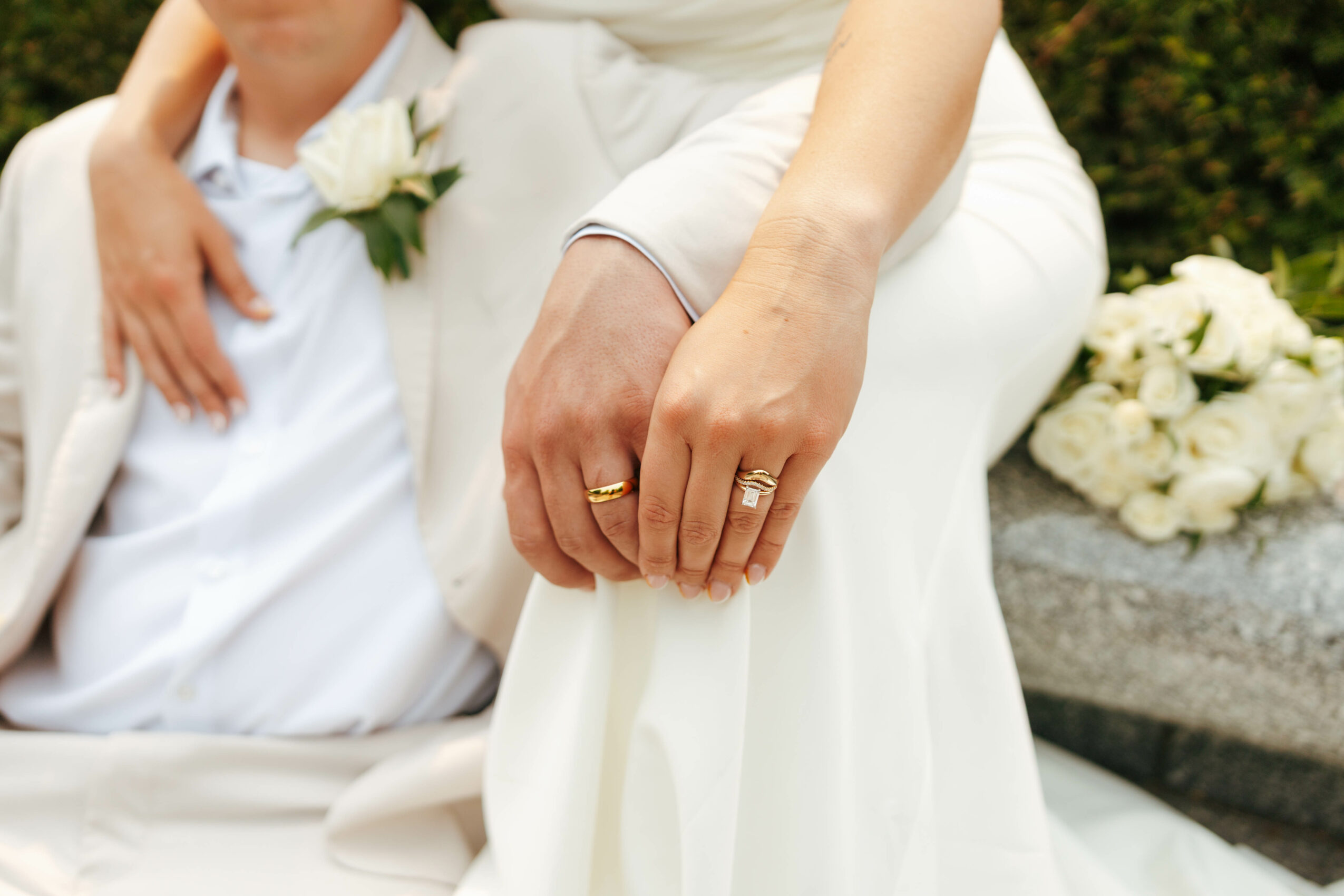 Close-up of a bride and groom's hands, with wedding rings, outdoors with flowers in background.