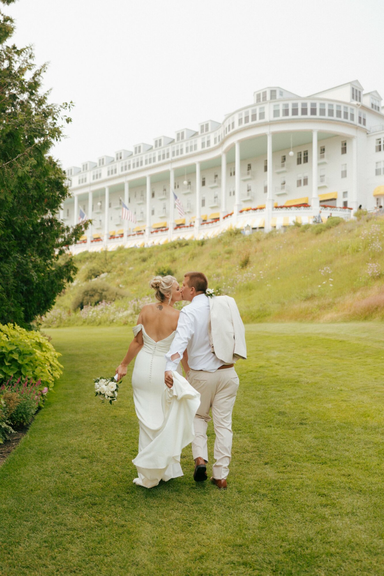 A bride and groom walk on a grassy area with a large white building in the background, holding hands and carrying flowers.