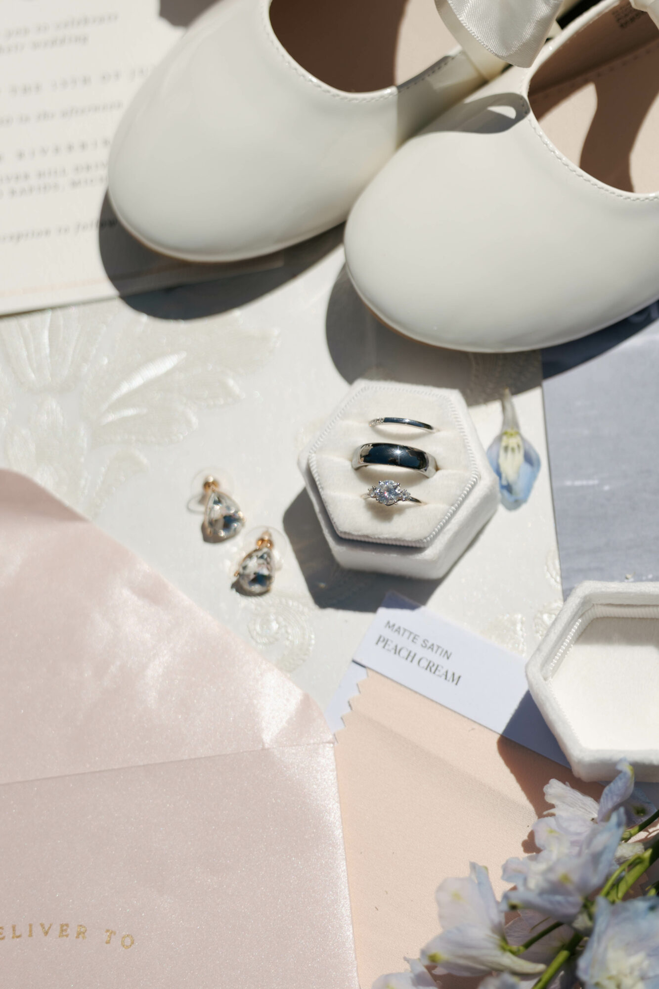 Pair of white shoes, jewelry in a box, and a pink cloth on a surface with a flower, with sunlight casting shadows.