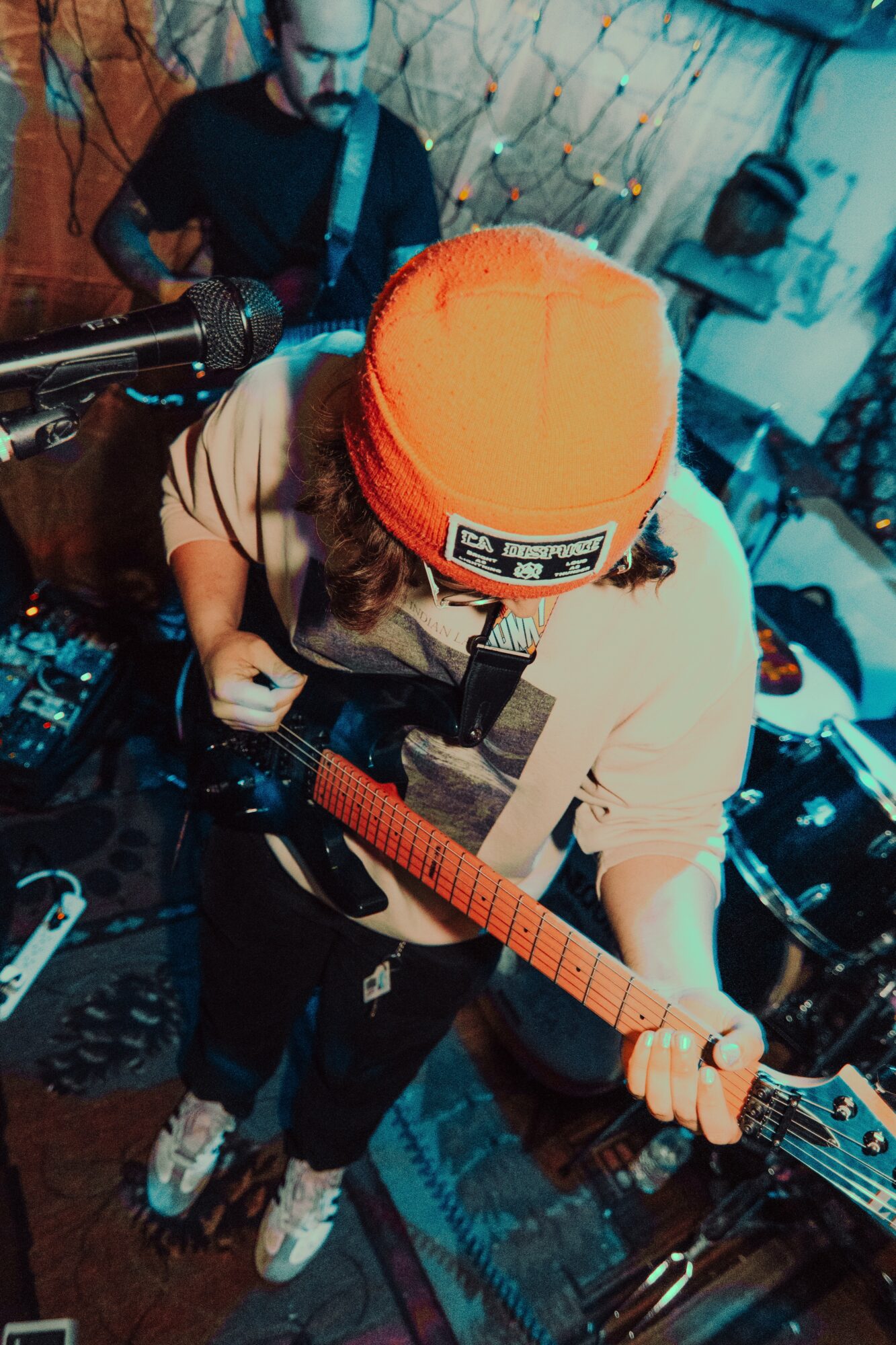 Person wearing orange beanie playing guitar, with a musician in the background, in a cozy indoor setting.