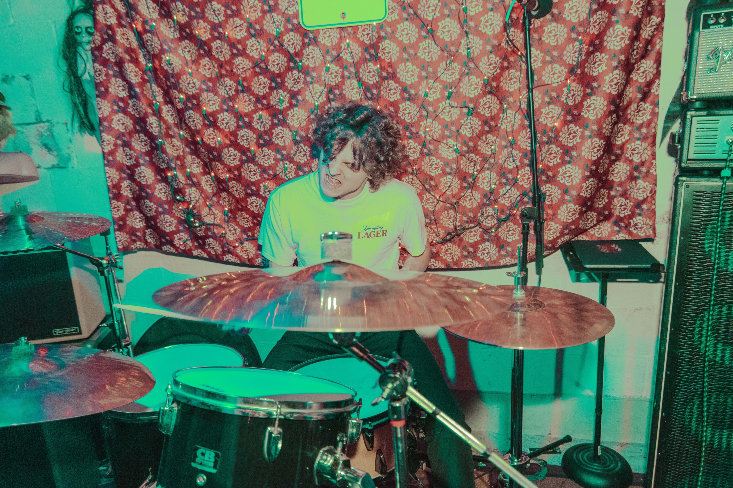 Person sitting behind a drum set, looking at a phone, with a patterned backdrop and audio equipment nearby.