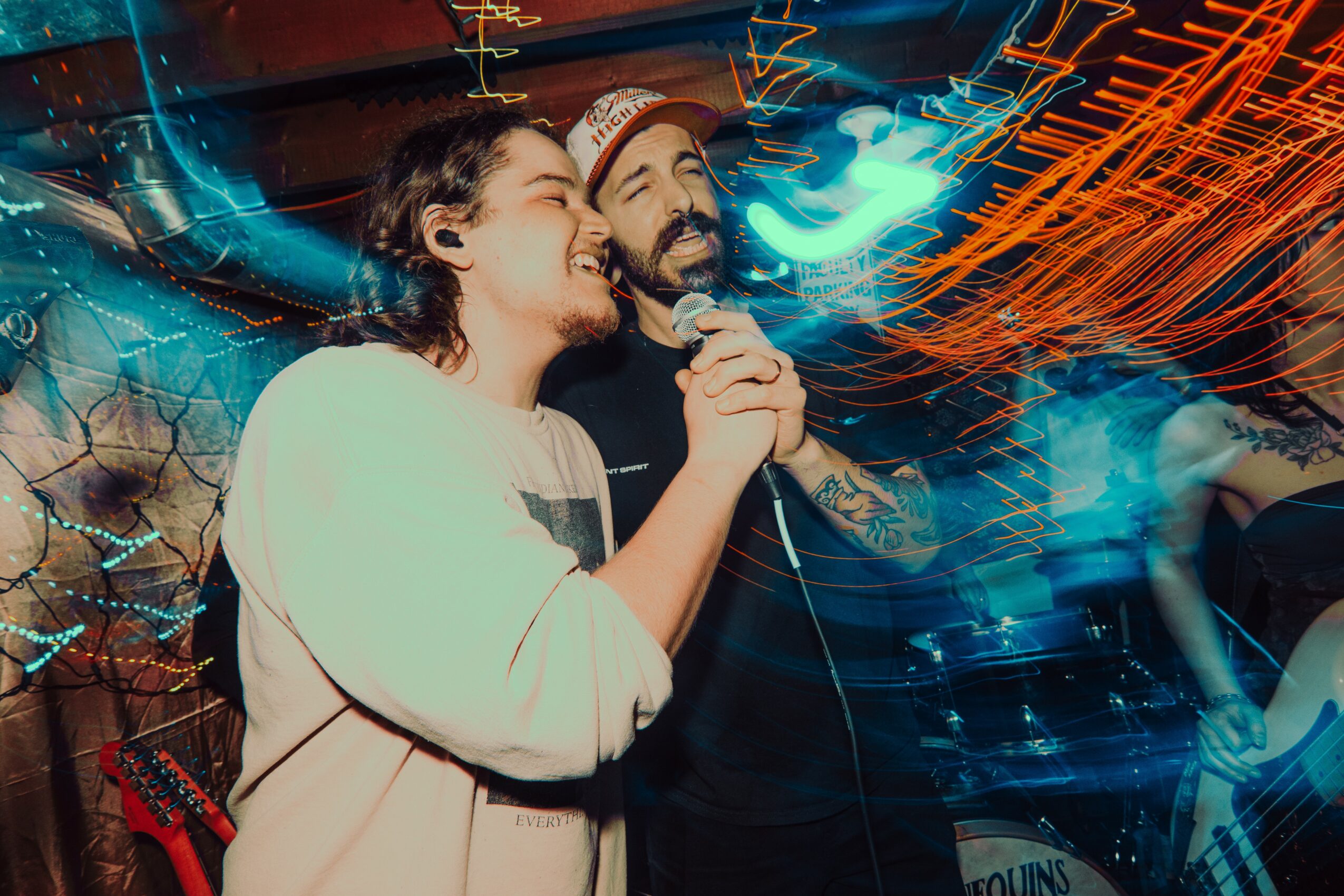 Two people singing into a microphone at a lively event with colorful lights and motion blur.