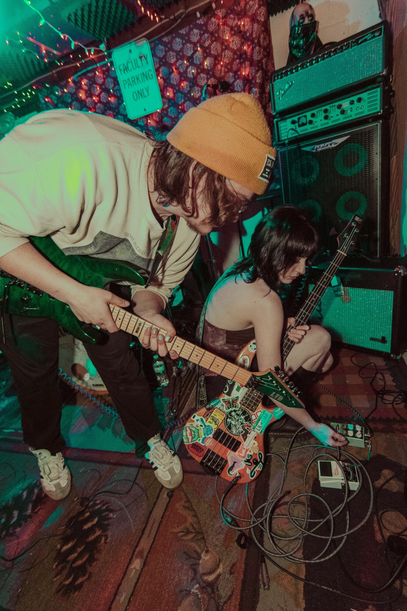 Two people playing electric guitars in a room with amplifiers and colorful decorations.
