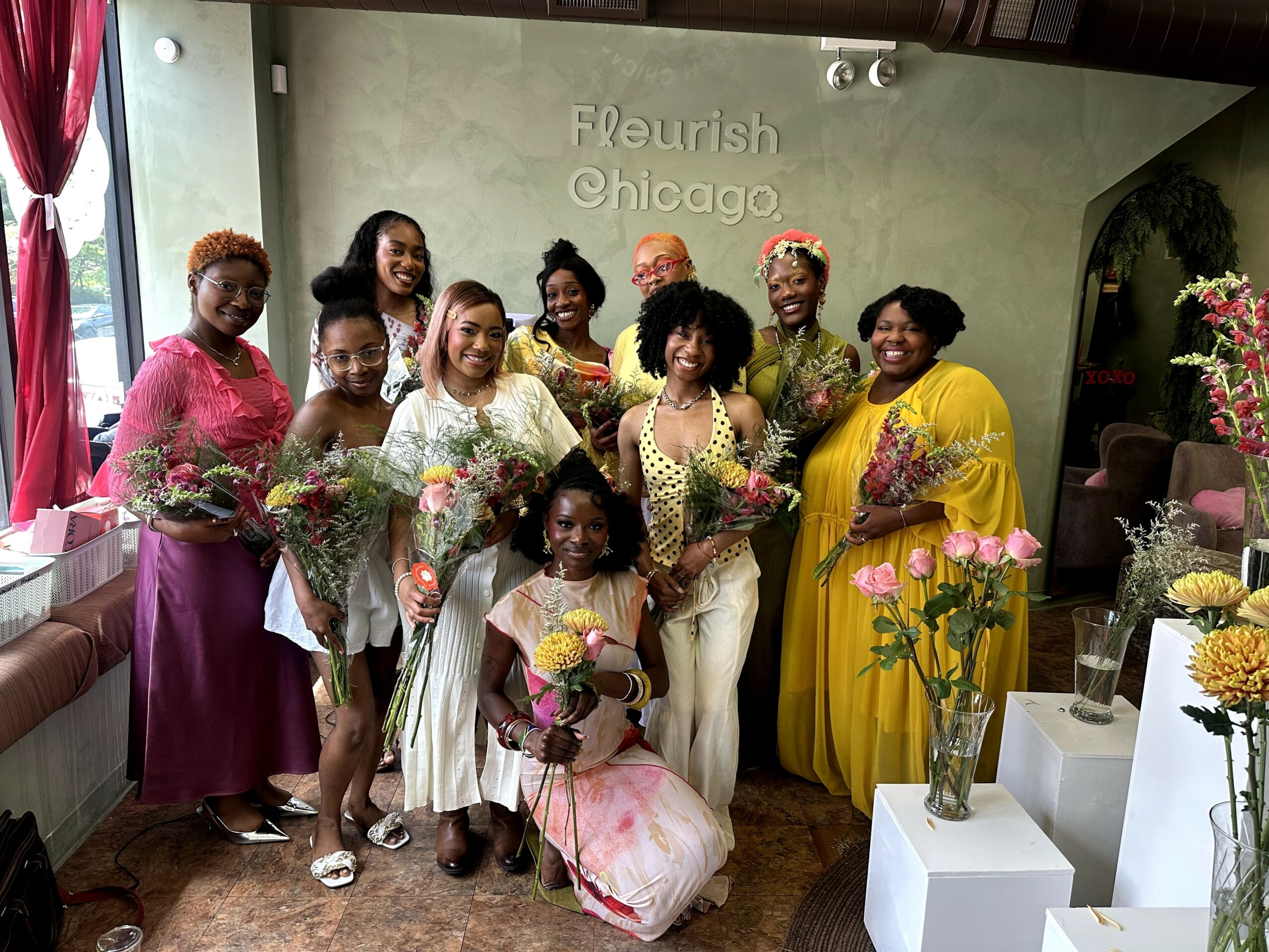 Group of women and girls holding flowers in a room with floral arrangements and a green wall, some wearing colorful dresses.