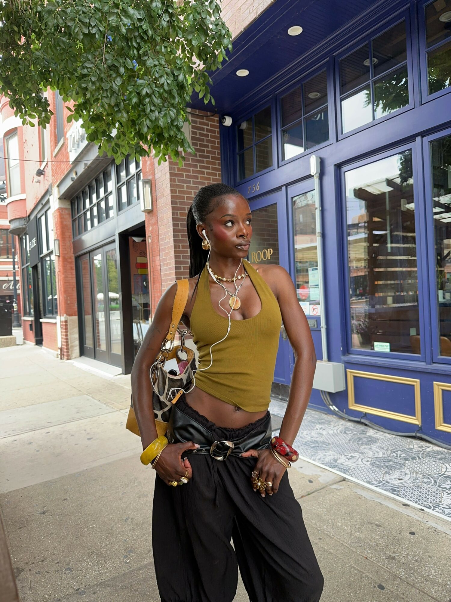Woman standing outside a building with blue and gold doors, wearing a yellow top and black pants, holding a bag.