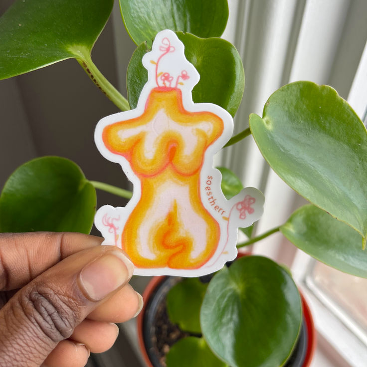 Person holding a candle-shaped sticker with orange and yellow colors, in front of a green plant near a window.