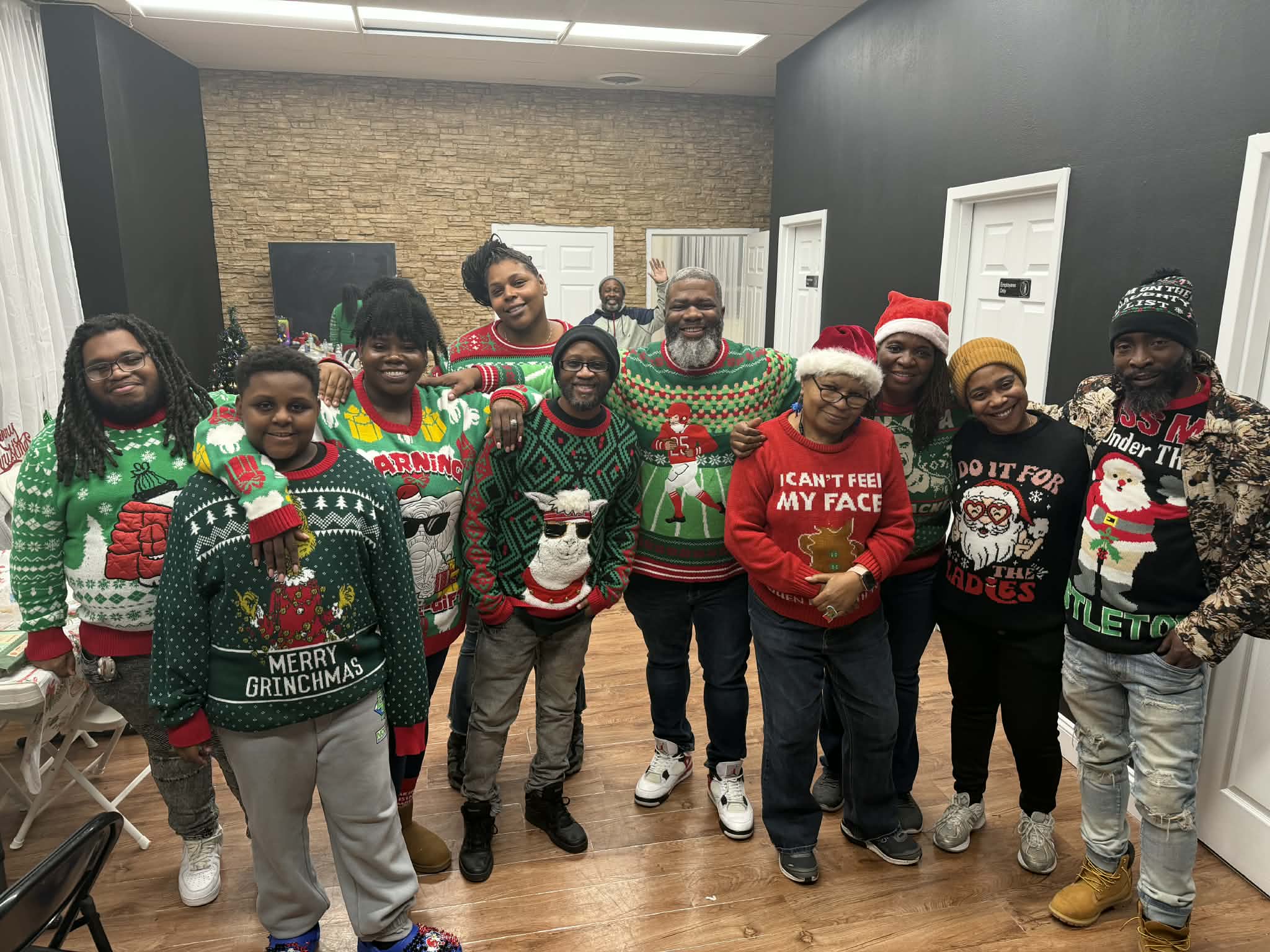 Group of people wearing Christmas sweaters and hats, standing indoors with a brick wall and whiteboard behind them.