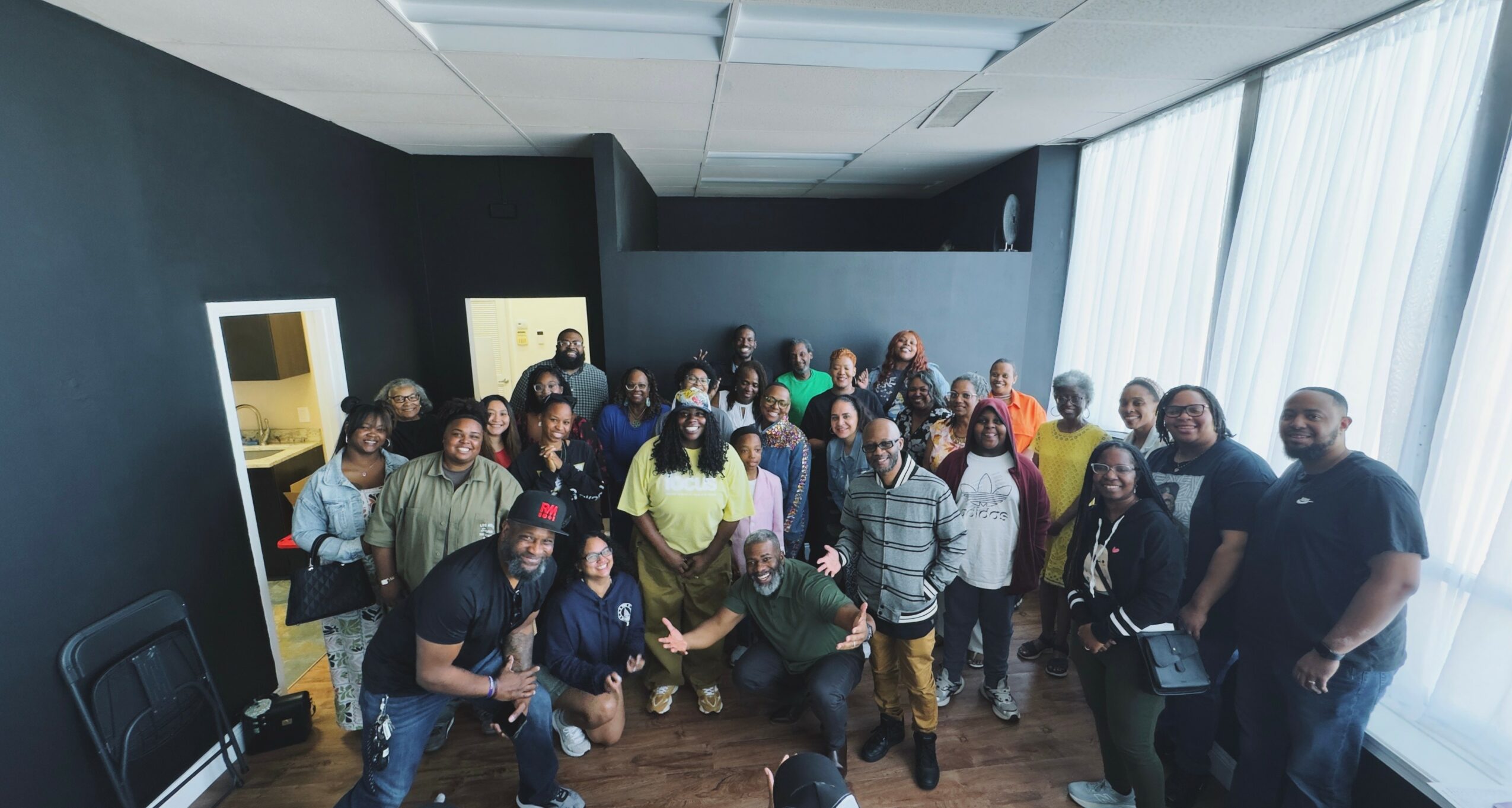 Group of people gathered in a room with large windows and dark walls, posing for a photo.