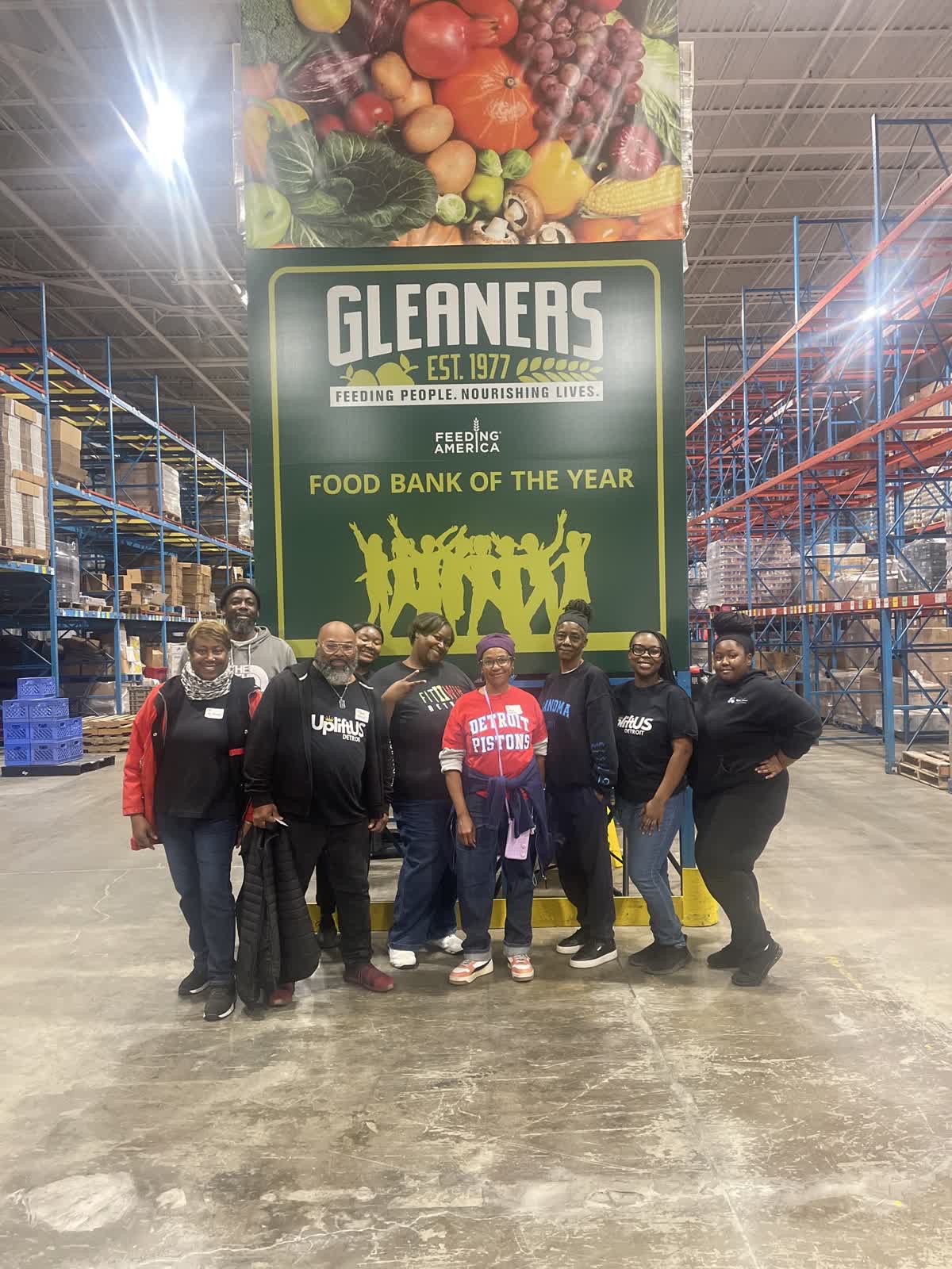 Group of six people standing in front of a large sign inside a warehouse, with shelves and products visible in the background.