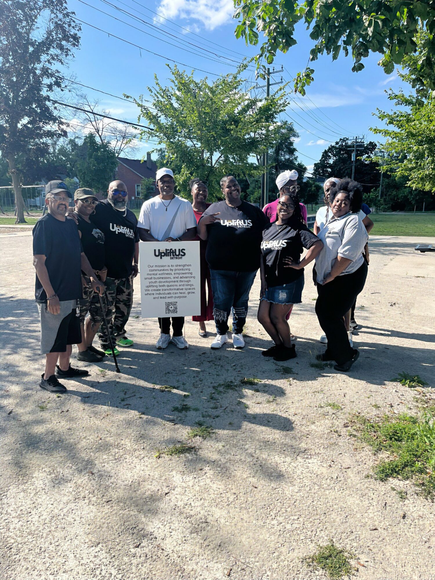 Group of people standing outdoors on a sunny day, some wearing helmets, with trees and a sign in the background.
