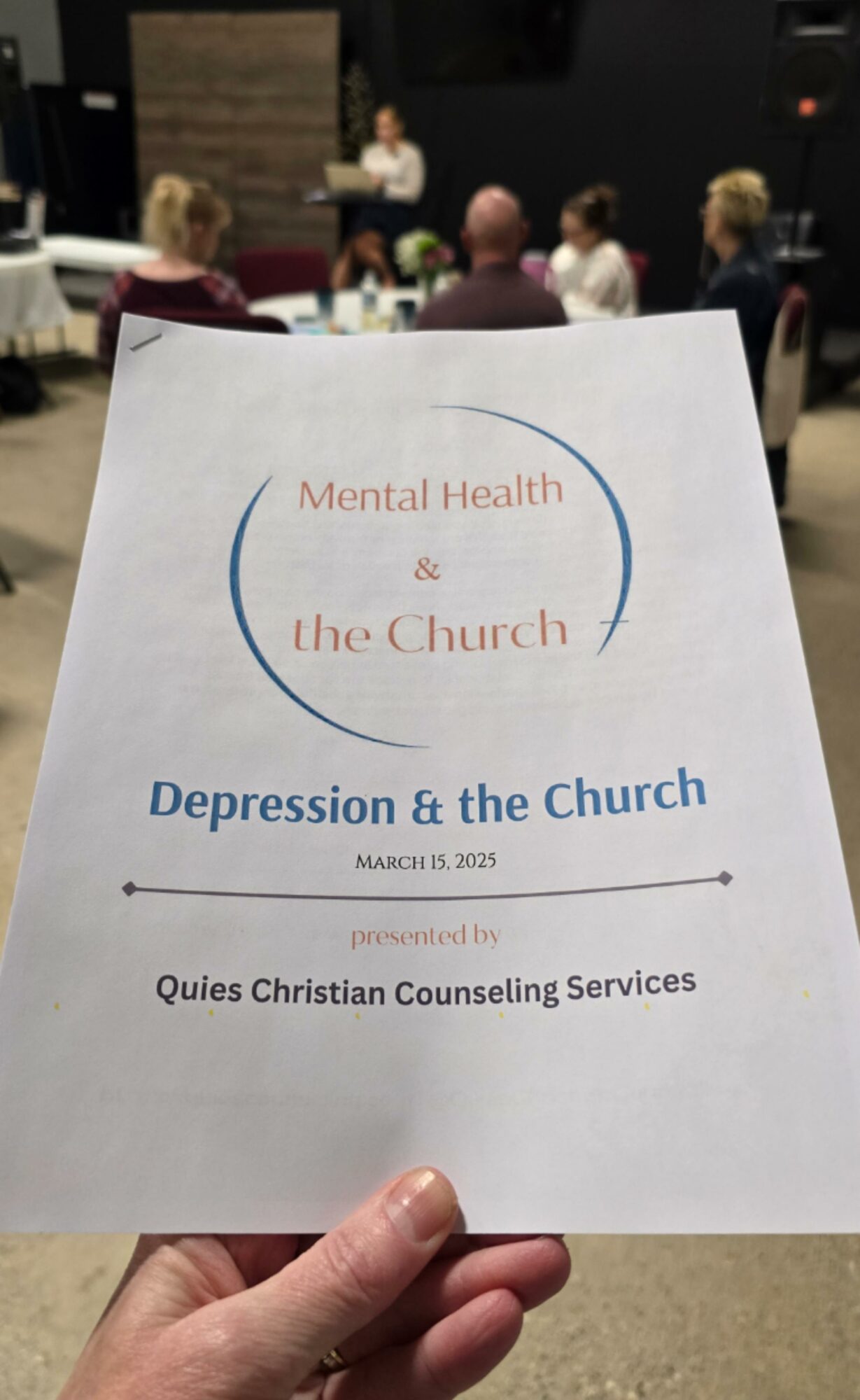 Hand holding a flyer titled 'Mental Health & the Church' with a group of people in a room in the background.