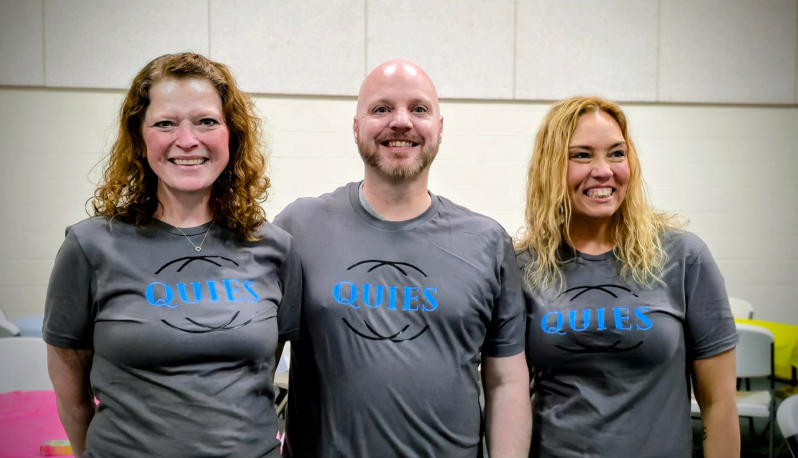 Three smiling people standing side by side in front of a plain wall, wearing matching gray shirts with blue text.