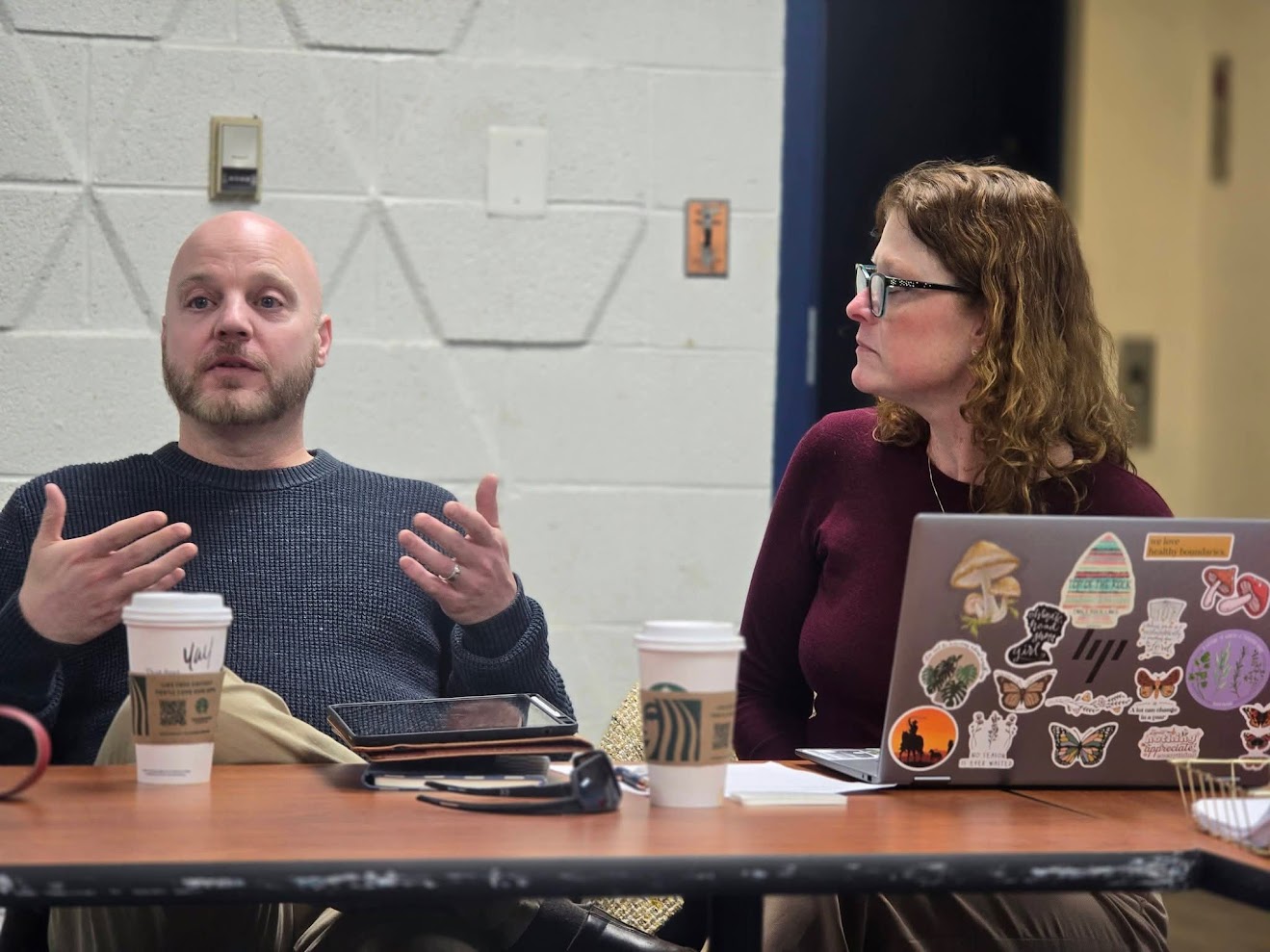 Two people sit at a table engaged in conversation, with coffee cups and a laptop decorated with stickers in front of them.