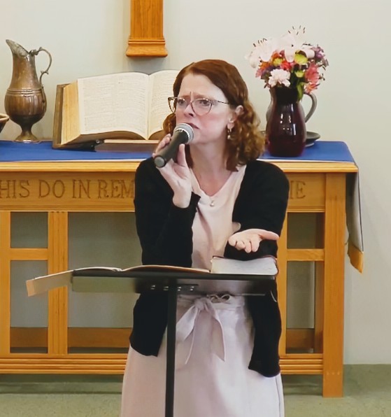Woman with glasses speaking into a microphone in front of a wooden table with a book, vase, and flowers.