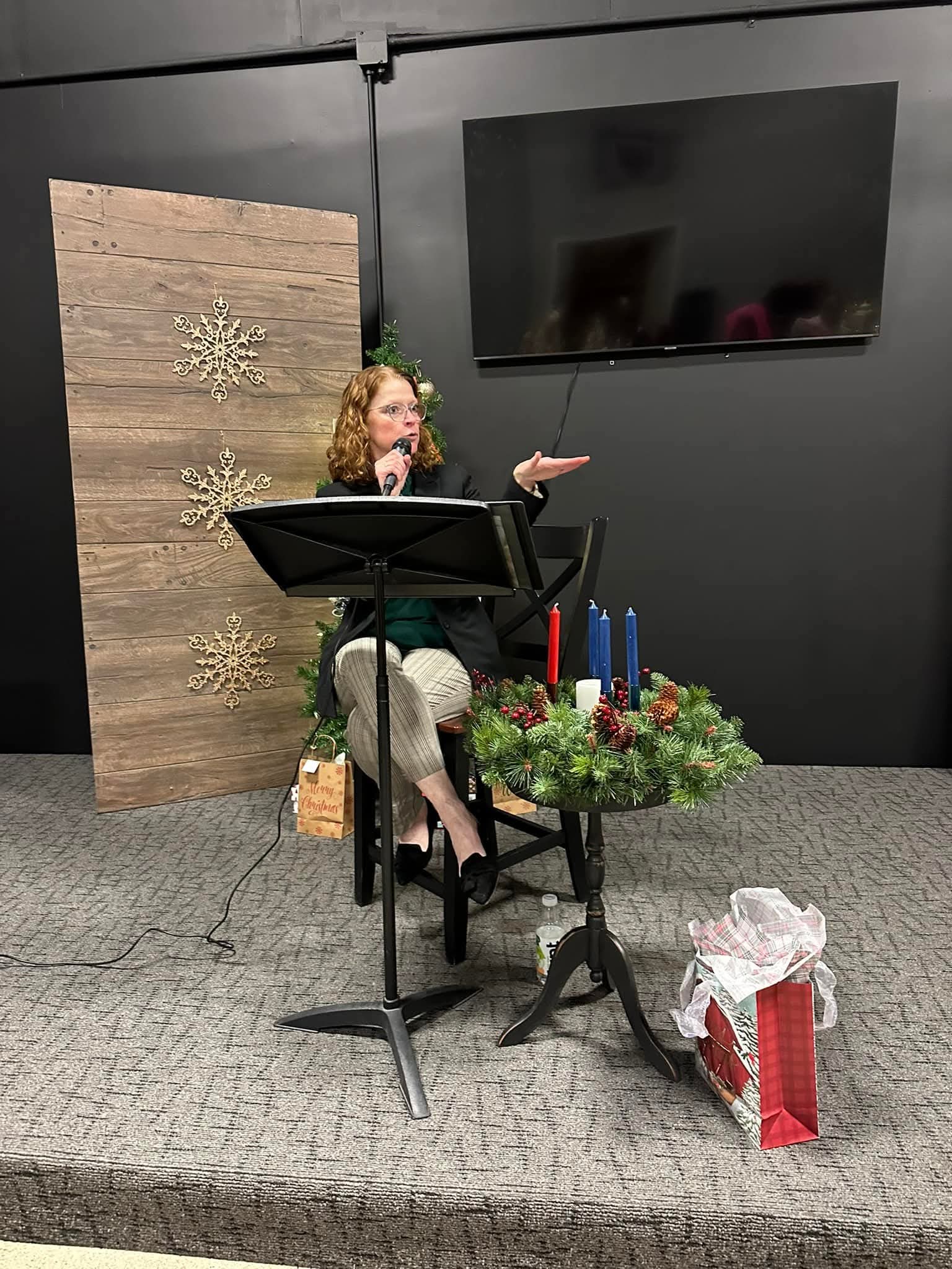 Woman sitting at a music stand, gesturing with her right hand, with a decorated table and a gift bag nearby.