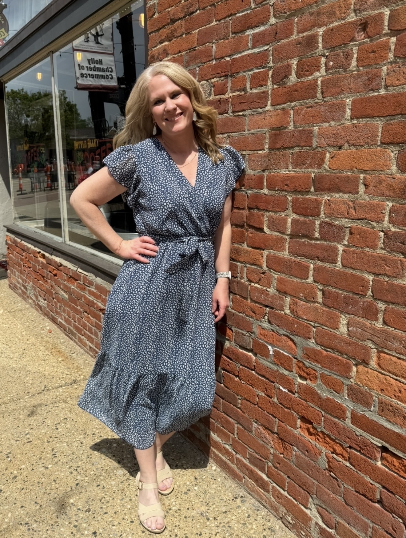Woman with blonde hair in a blue patterned dress standing outdoors against a brick wall, smiling, with a store window nearby.