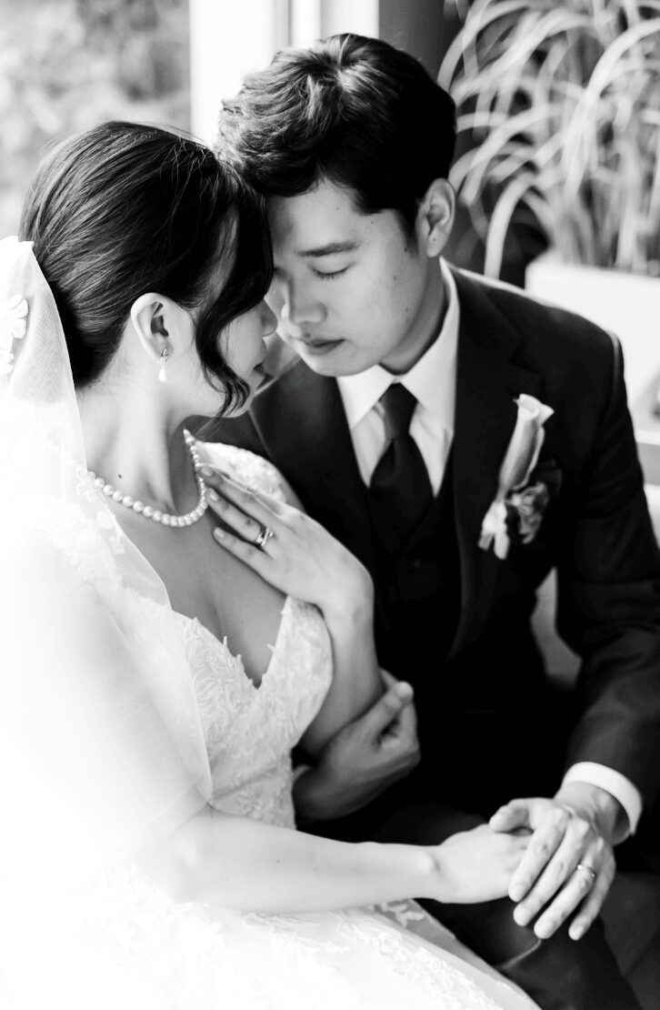 A bride and groom sit close, touching foreheads and holding hands, in a black and white wedding photo.