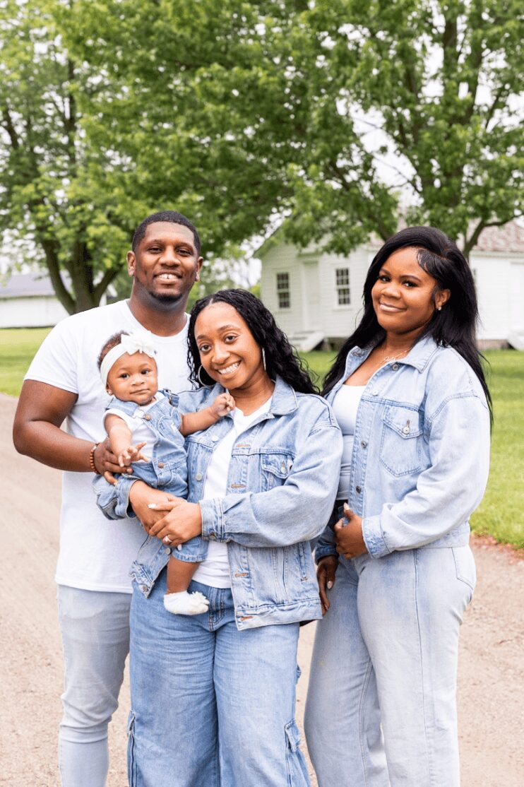 Group of four people outdoors, smiling, with trees and a house in the background.