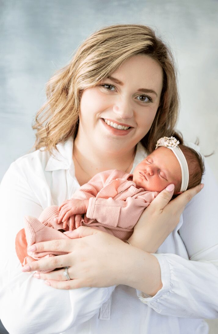 Woman holding a newborn baby, smiling, against a light background.