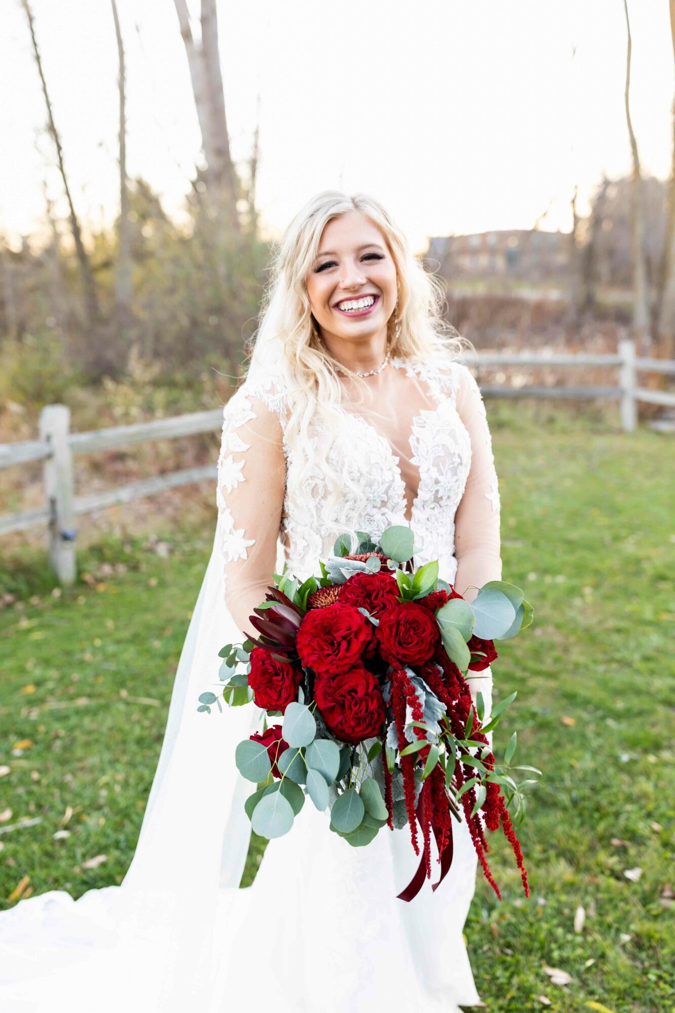 Smiling bride in a white wedding dress holding a large red and green bouquet outdoors.