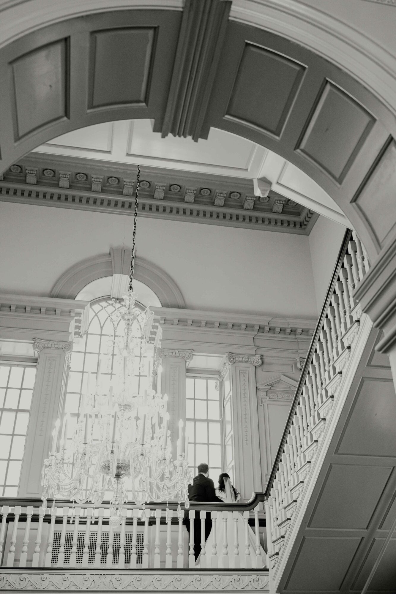 Interior view of a grand hall with a large chandelier, high windows, and a staircase, with two people standing near the railing.