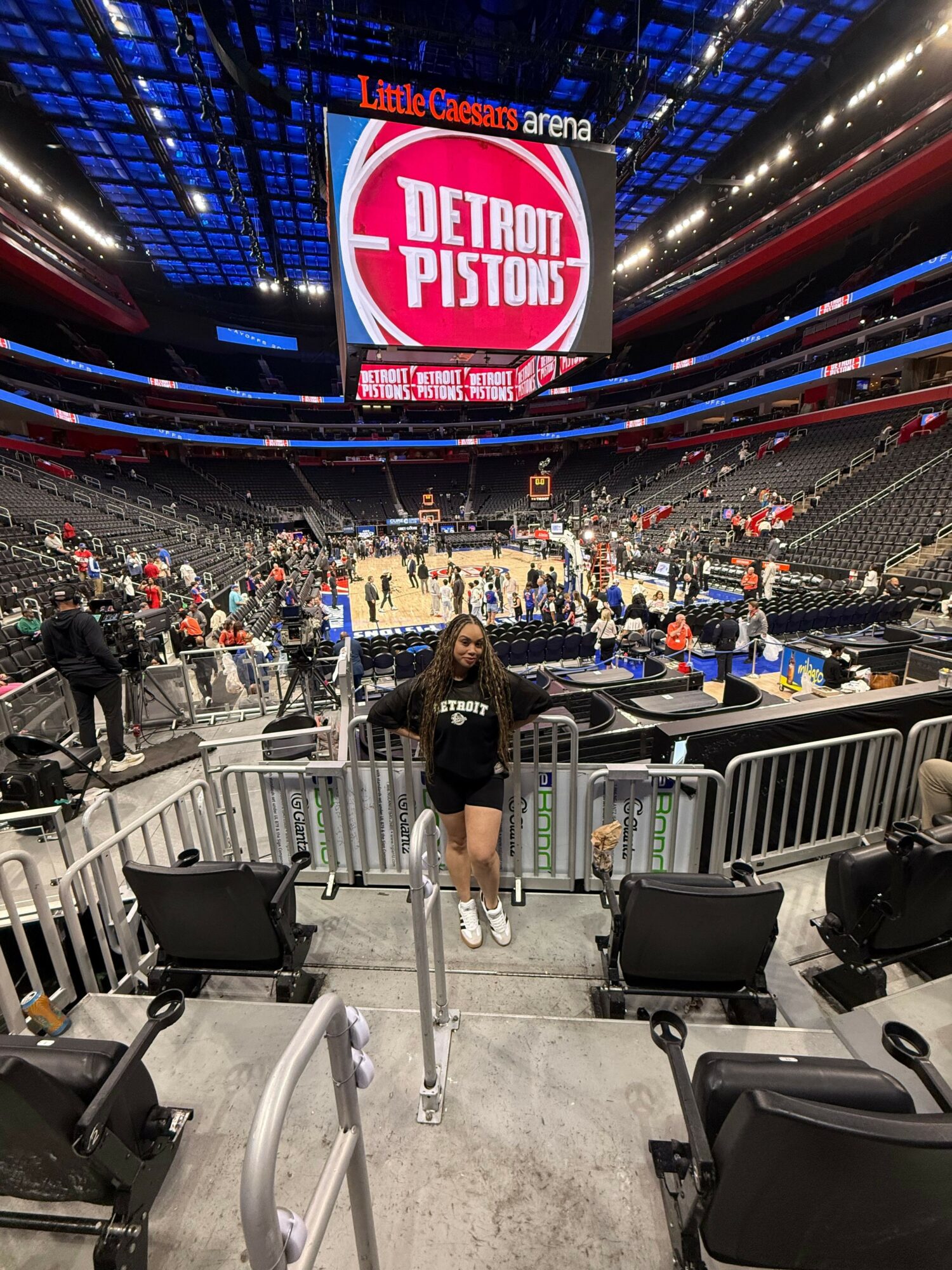 Person standing in a large indoor stadium with a crowd, electronic screens, and a sign reading 'Patriots Playoff' in the background.